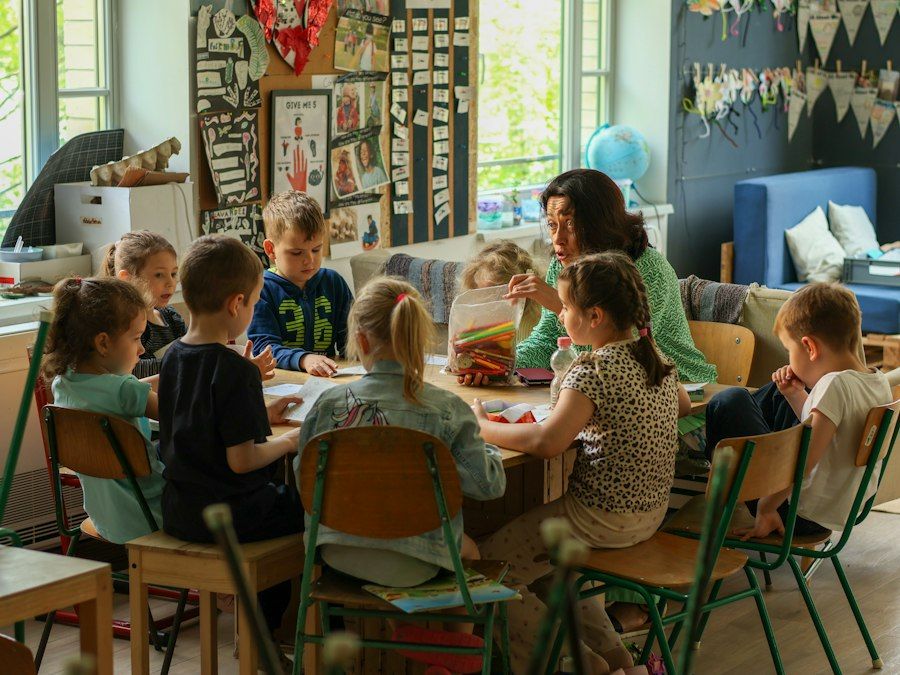 Odgajateljica sjedi za stolom okružena djecom koja se igraju i uče